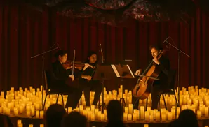 A string trio performs in front of a red curtain, surrounded by a sea of glowing candles.