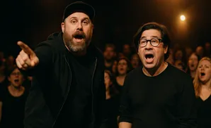 Two men sing with mouths open, one pointing forward, in front of a blurred chorus.