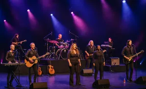 A band performs on stage with purple and blue lights. The band members play guitars, drums, and keyboards.