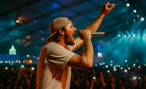A male singer in a cap and t-shirt sings into a microphone on stage with a cheering crowd.