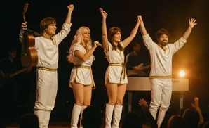 Four performers in white, gold-trimmed costumes, two women singing, two men playing instruments on stage.