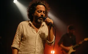 Singer with curly hair and beard sings into a microphone under a spotlight, another musician plays guitar in the background.