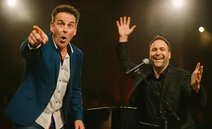 A man in a blue jacket points while another in a black suit raises his hand by a piano.