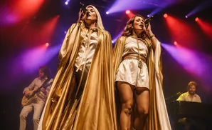 Two women in gold capes and white outfits sing into microphones on a stage with purple and red lights.