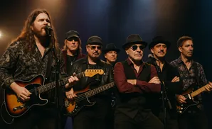 A band of six men playing guitars and singing on stage.