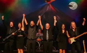 A band of six people, dressed in black, pose with their hands raised in celebration on stage with a volcano backdrop.