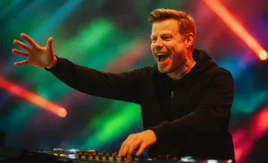 A joyful DJ with blonde hair and a beard in a black hoodie, reaching out with one hand while mixing on a DJ board, illuminated by colorful stage lights.