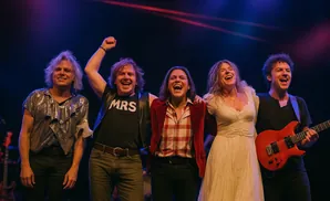 Band members on stage, smiling and laughing. Two men in the front wear sparkly shirts, one with 