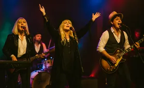 A band performs on stage with colorful lighting. The lead singer in the center has her arms raised.