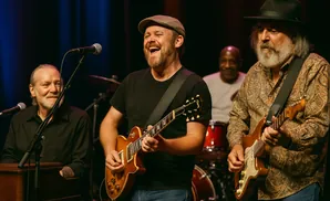 A band performs on stage with a vocalist and guitarist laughing in the center.
