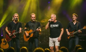 A band performs on stage with yellow and purple spotlights. One man sings into a microphone, another plays guitar, and a third plays bass guitar.