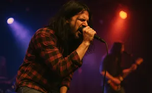A man with long hair sings into a microphone on a dimly lit stage, wearing a red and black plaid shirt.