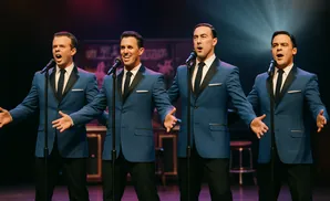 Four singers in matching blue suits and ties perform on stage with microphones.
