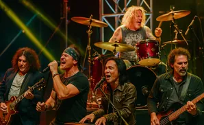 A rock band performs on stage with spotlights, the singer is in the foreground singing into a microphone.