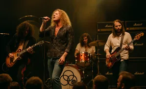 A rock band performs on stage, with a singer in the foreground and guitarists and a drummer in the background.