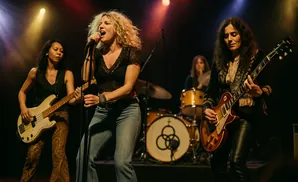 A musician with wavy blonde hair stands on a stage, back to the camera, facing a massive crowd at a concert.