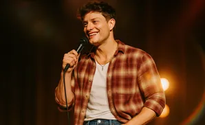 A man in a plaid shirt and jeans holds a microphone and smiles.