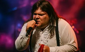 A male singer with long dark hair and a white ruffled shirt sings into a microphone, holding a red cloth.