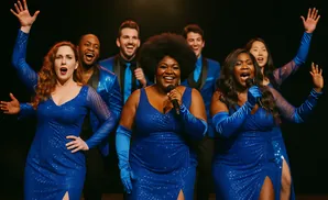 A diverse group of singers in matching blue sequined dresses and suits performs on stage.