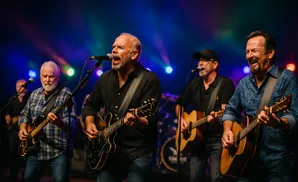 Four men singing and playing guitars on a stage with colorful lights in the background.