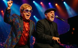 Elton John and Billy Joel perform together on stage with Elton in a sparkly jacket and Billy at the piano.
