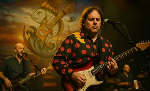 A band performs on stage, with a guitarist in a polka-dot shirt in the foreground.