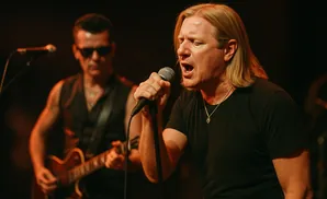 Singer with blonde hair and open mouth sings into a microphone, with a guitarist in sunglasses behind him.