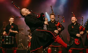 Bagpipers in black and red kilts perform on stage with drums and spotlights.