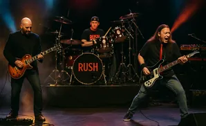 A three-person band performs on stage, with two guitarists and a drummer.