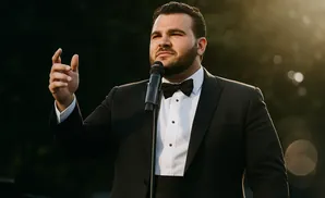 A man in a tuxedo sings into a microphone, gesturing with his left hand.