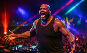 Shaquille O'Neal in a black tank top, shouting with arms open on a stage with colorful lights and a crowd.