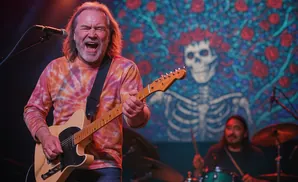 A man with a tie-dye shirt plays a guitar on stage with a skeleton and roses backdrop.