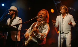A male musician in a shiny shirt and vest laughs while playing a guitar on stage.