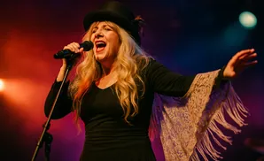 A woman in a black top hat sings into a microphone, wearing a black dress with a lace shawl with fringe.