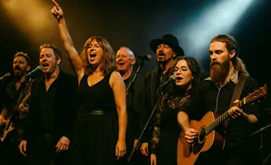 A band performs on stage under a spotlight, with a female singer in the foreground pointing upwards.