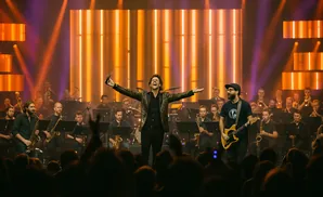 A conductor leads a band with a saxophonist and guitarist on stage, bathed in orange stage lights.