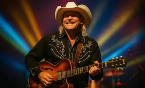 A smiling man in a cowboy hat plays an acoustic guitar on stage under colorful lights.