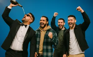 Four men celebrate joyfully. One man in a tuxedo pours champagne, while the others raise their fists in excitement.