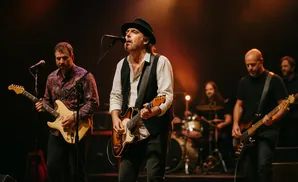A band performs on stage with the lead singer in a hat and vest singing into a microphone.