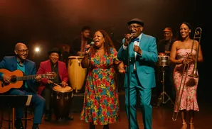 A band performs on stage with singers, a guitarist, conga drums, and a trombonist.