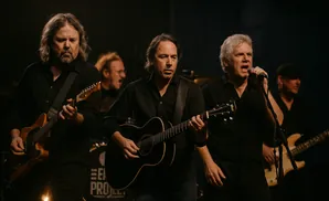A band of four men in black shirts perform on stage, playing guitars and singing into microphones.