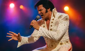 Elvis Presley impersonator singing into a microphone on stage in a sparkling white jumpsuit.