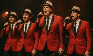 Four men in red suits and fedoras sing into microphones on a dark stage.