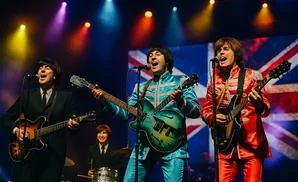 A Beatles tribute band performs on stage, with three members playing guitars and singing, backed by a Union Jack flag.