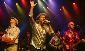 A band performs on stage under colorful spotlights. The lead singer, wearing a shiny silver shirt, holds a microphone and has his arm raised.