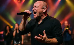A male singer in a black shirt performs with a microphone, with a blurred band in the background.