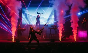 A band performs on stage with smoke, lasers, and a screen showing a mountain scene.