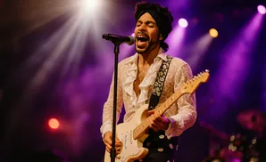 A male singer in a sheer ruffled shirt and polka dot pants plays a white electric guitar under purple stage lights.
