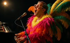 Elton John in a flamboyant feather costume sings into a microphone at a piano.