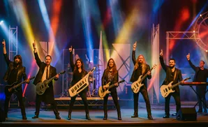 A band of six musicians performing on stage with guitars and keyboards, illuminated by colorful stage lights.